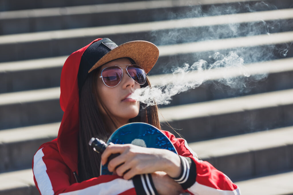 Vaping girl. Young hipster woman vape e-cig. Pretty young female in black hat, red clothing vape ecig, vaping device at the sunset. Toned image. Closeup. Hip-hop style.