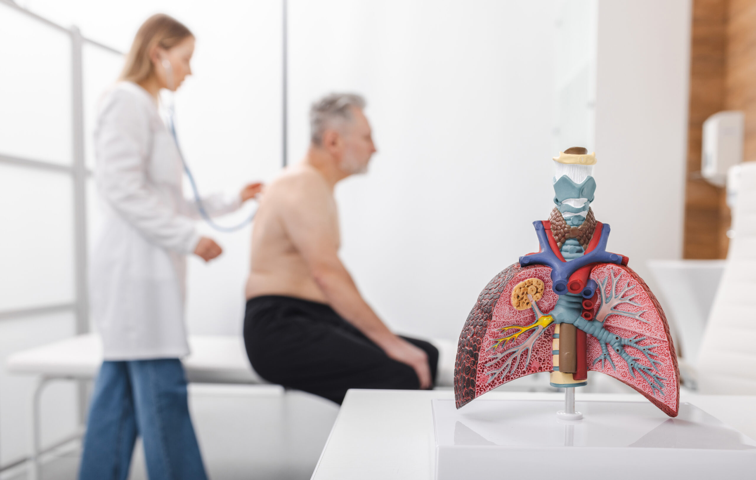 Senior patient sits on examination table while a doctor examines his lungs using a stethoscope in a medical clinic setting